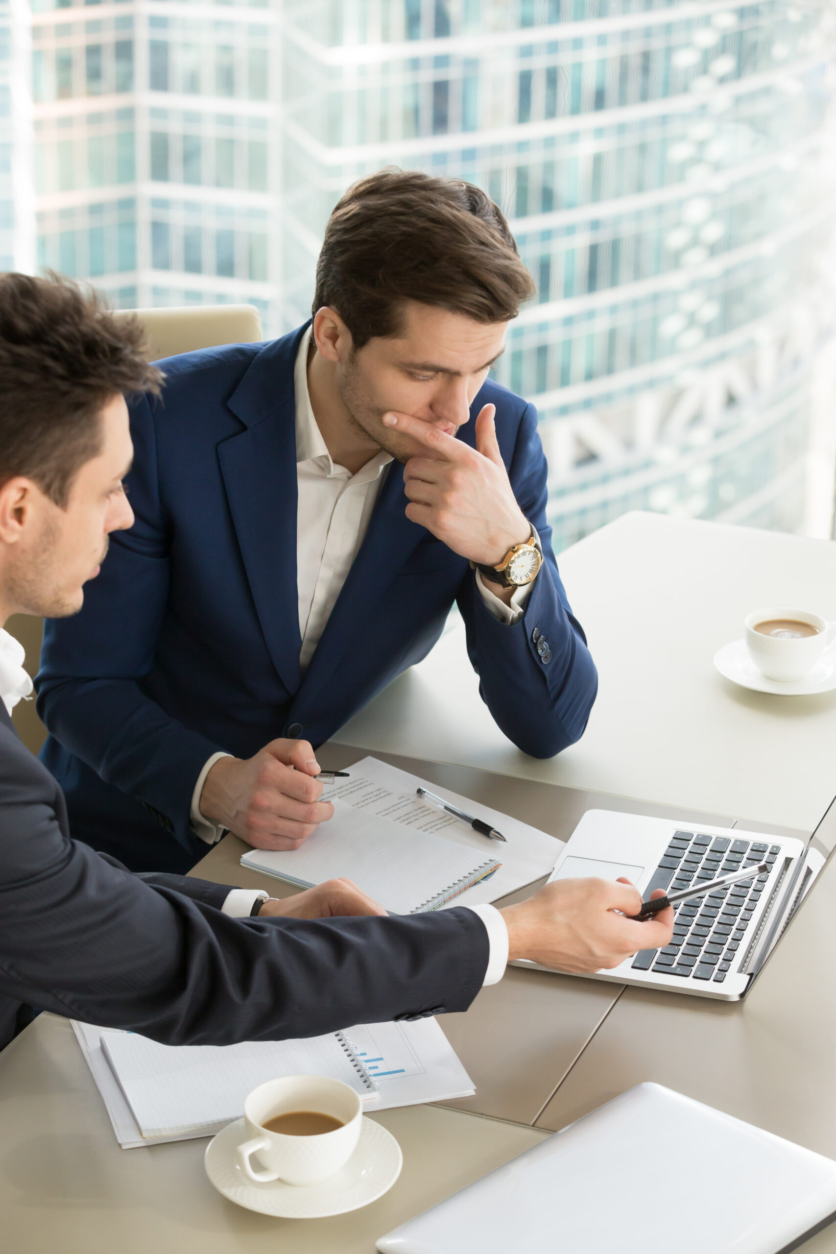 Deux hommes d'affaires en costume sont assis à un bureau avec un ordinateur portable, des documents et des tasses à café. L'un pointe l'écran de l'ordinateur tandis que l'autre a l'air pensif, tous deux entourés de grandes fenêtres.