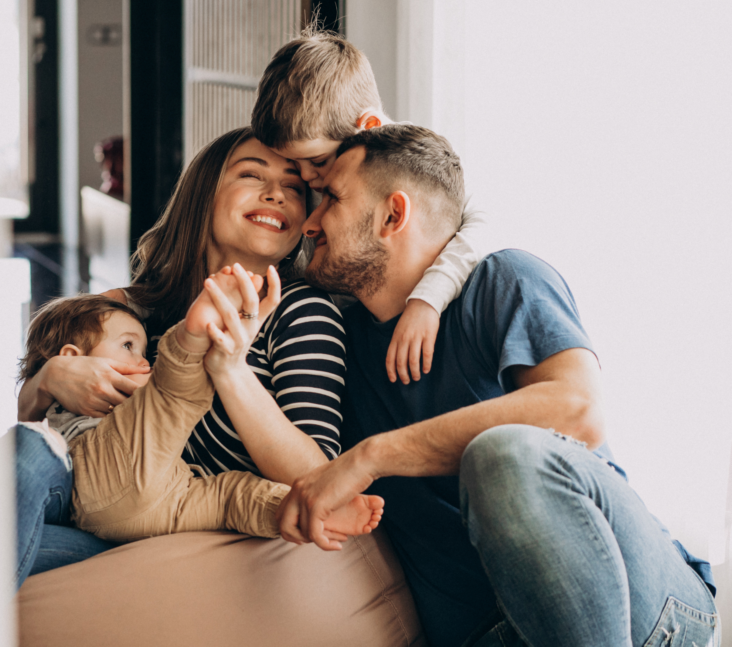 Une famille heureuse de quatre personnes est assise à l'intérieur, les bras croisés, souriant et se faisant des câlins. La mère, tenant un jeune enfant dans ses bras, rit tandis que le père la serre dans ses bras et qu'un enfant plus âgé s'appuie affectueusement sur ses deux parents.